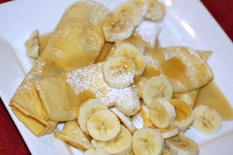 Pile of crepes with powdered sugar, bananas, and syrup.