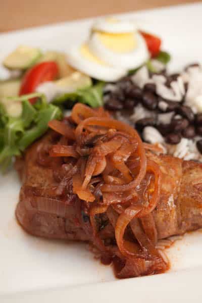 A close-up of a pork chop topped with onions with rice, beans, and a salad.