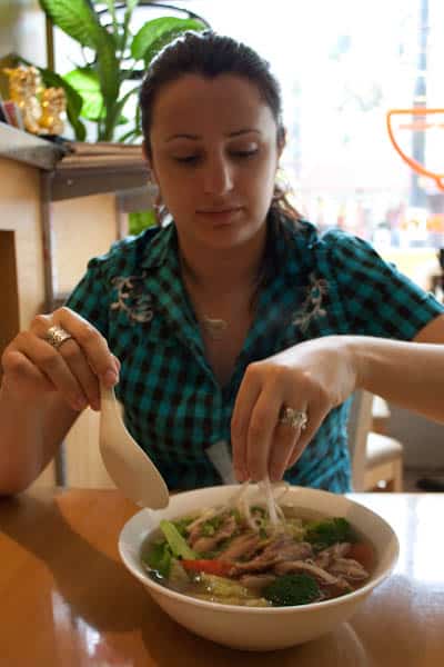 A woman sitting at a table sprinkling bean sprouts on pho.