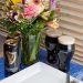 A vase of flowers on a table with beer and an empty plate.