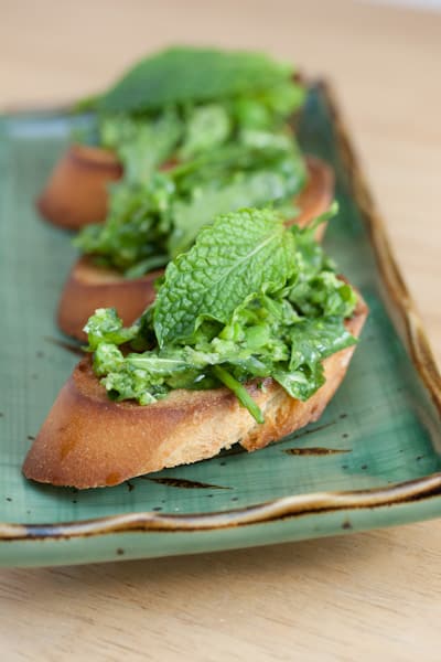 A close-up of crostini topped with fava bean, arugula, and mint.