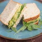 A sandwich that is filled with an avocado and chickpea mixture and baby arugula, sliced in half on a blue plate.