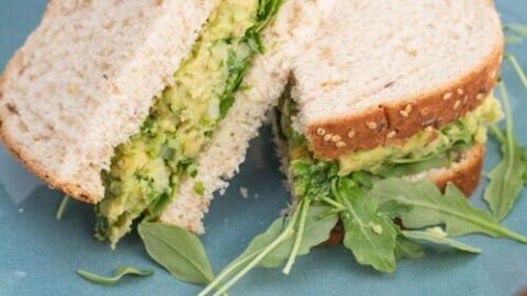 A sandwich that is filled with an avocado and chickpea mixture and baby arugula, sliced in half on a blue plate.