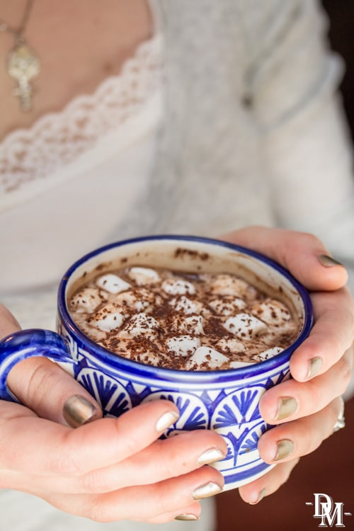 Holding a mug of hot chocolate with chocolate shavings on top.