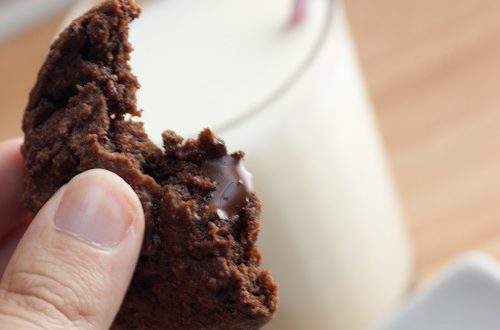 Hand holding a chocolate cookie with melted chocolate chips and a glass of milk.