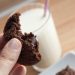 Hand holding a chocolate cookie with melted chocolate chips and a glass of milk.