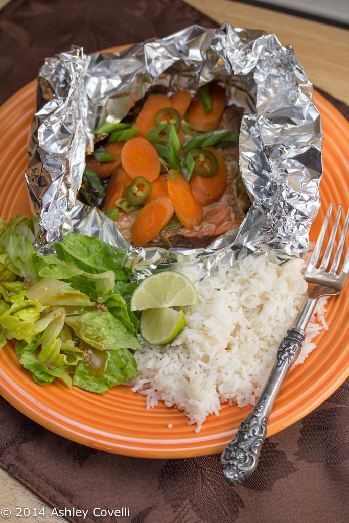 Salmon and carrots in a foil packet on a plate with rice and green onions.