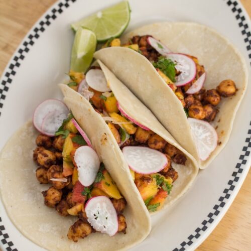 Overhead view of a plate with 3 Spiced Chickpea Tacos with Cilantro-Peach Relish and lime wedges.