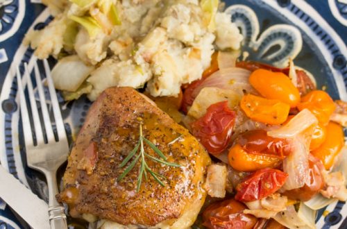 Overhead view of a plate with chicken thighs covered in a pan sauce and fresh rosemary next to colorful tomatoes and mashed potatoes.