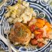 Overhead view of a plate with chicken thighs covered in a pan sauce and fresh rosemary next to colorful tomatoes and mashed potatoes.