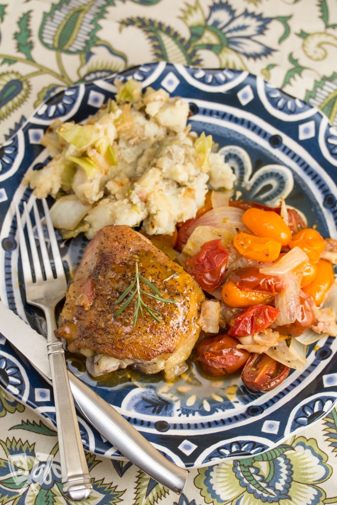 Overhead view of a plate with chicken thighs covered in a pan sauce and fresh rosemary next to colorful tomatoes and mashed potatoes.