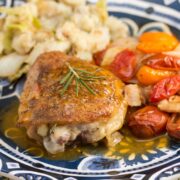 ¾ view of a plate with chicken thighs covered in a pan sauce and fresh rosemary next to colorful tomatoes and mashed potatoes.