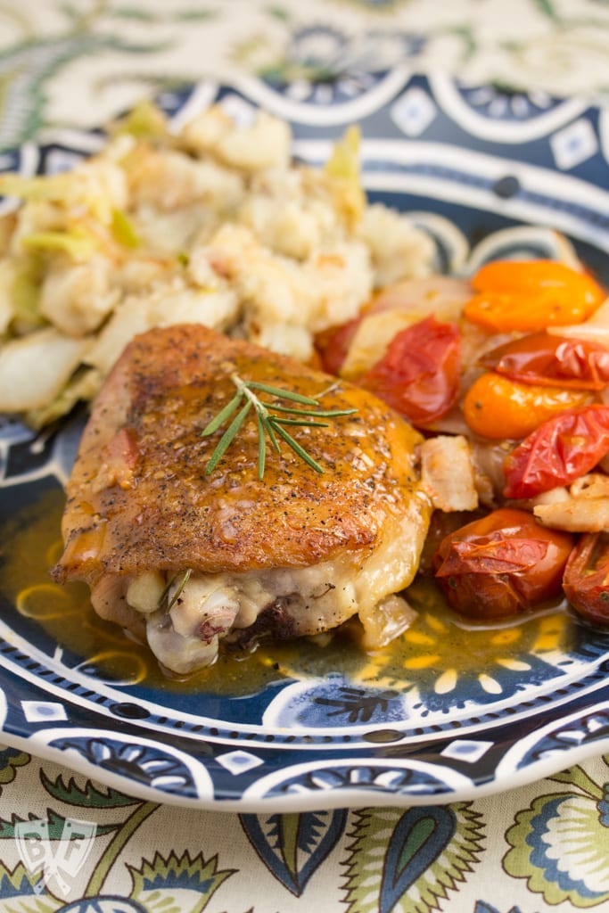 3/4 view of a plate with chicken thighs covered in a pan sauce and fresh rosemary next to colorful tomatoes and mashed potatoes.