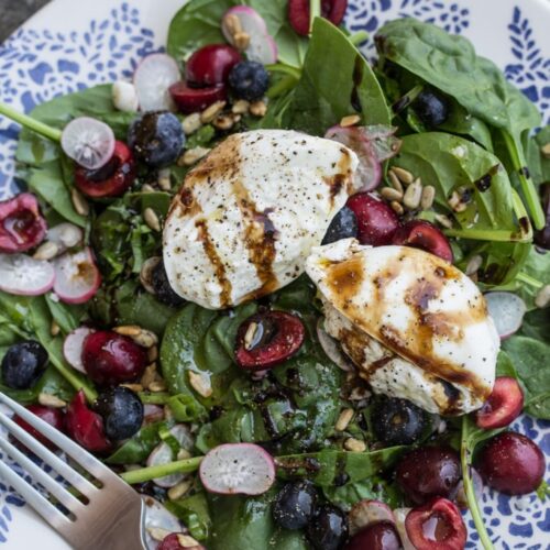 Overhead view of a plate of salad topped with fruit, vegetables, and burrata cheese drizzled with aged balsamic.
