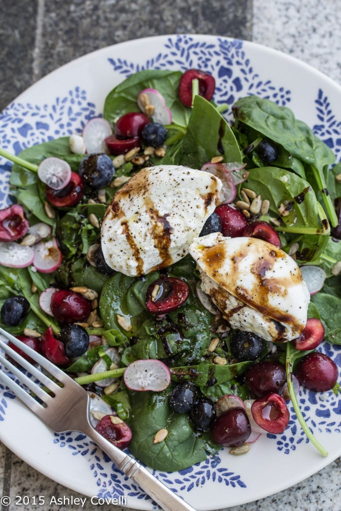 Overhead view of a plate of salad topped with fruit, vegetables, and burrata cheese drizzled with aged balsamic.