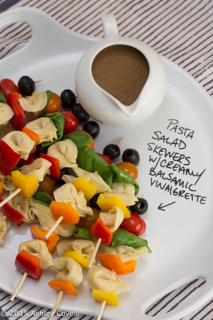 Skewers of pasta salad ingredients next to a pitcher of balsamic vinaigrette.