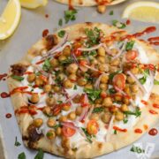 Overhead view of 2 flatbreads covered in tzatziki and topped with falafel-spiced chickpeas garnished with fresh herbs, lemon wedges, and sriracha sauce.