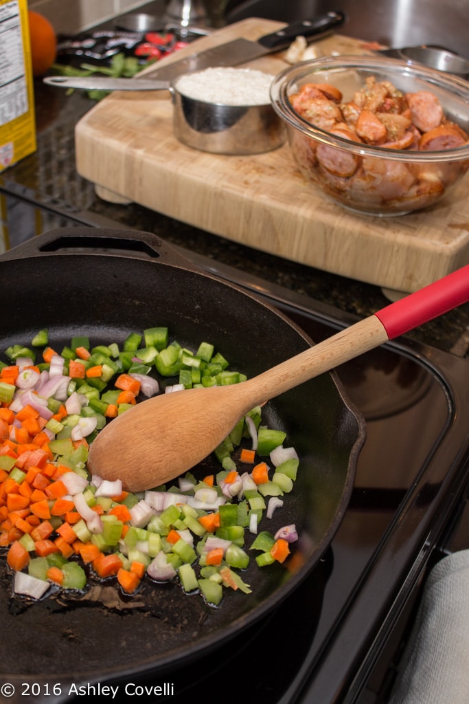 Stirring veggies in a cast iron skillet with sausage and rice in the background.