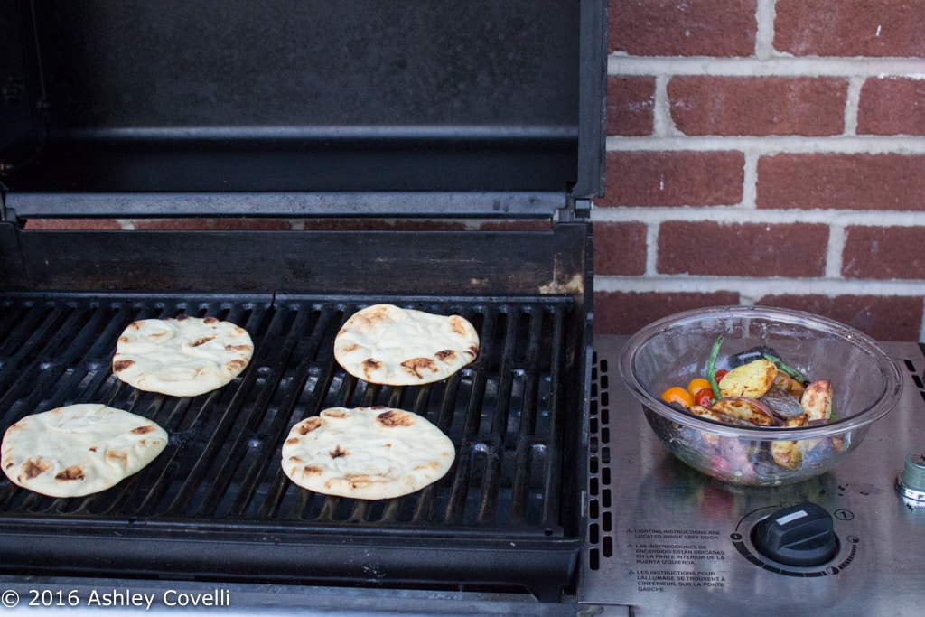 A grill with mini naan breads on top and a bowl of grilled veggies off to the side.