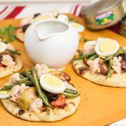 3 mini naan breads topped with grilled Niçoise salad ingredients along with a pitcher of vinaigrette and tuna cans in the background.