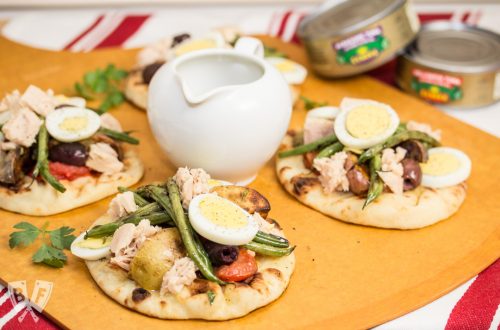 3 mini naan breads topped with grilled Niçoise salad ingredients along with a pitcher of vinaigrette and tuna cans in the background.