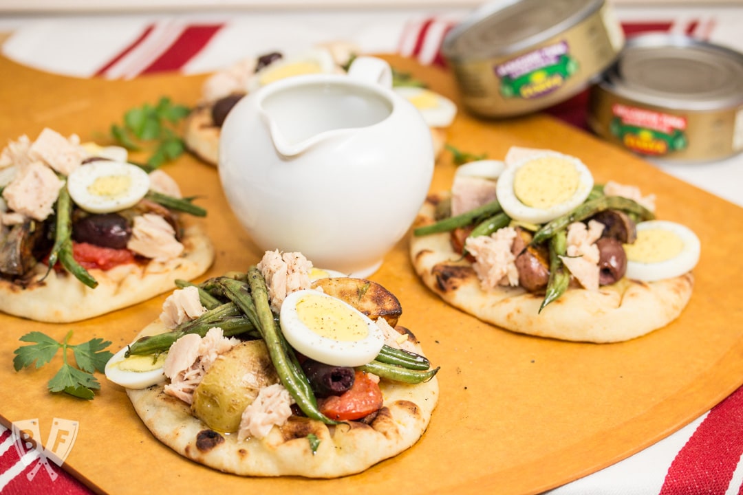 3 mini naan breads topped with grilled Niçoise salad ingredients along with a pitcher of vinaigrette and tuna cans in the background.