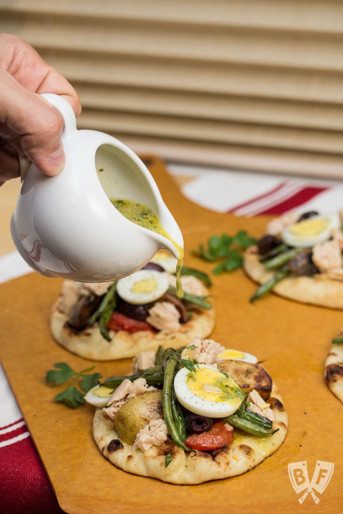 Pouring vinaigrette over a tray of mini naan breads topped with grilled Niçoise salads.