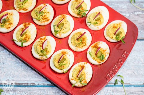 A red platter full of beautifully piped deviled eggs garnished with radishes and pea shoots.