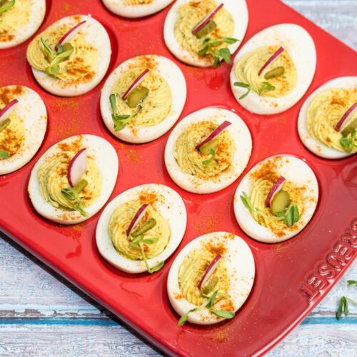 A red platter full of beautifully piped deviled eggs garnished with radishes and pea shoots.