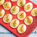 A red platter full of beautifully piped deviled eggs garnished with radishes and pea shoots.