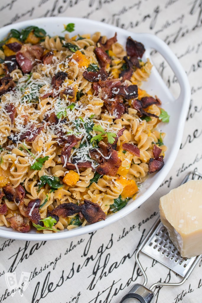 Bowl of red lentil fusilli with bacon, butternut squash, and spinach topped with Parmesan cheese.