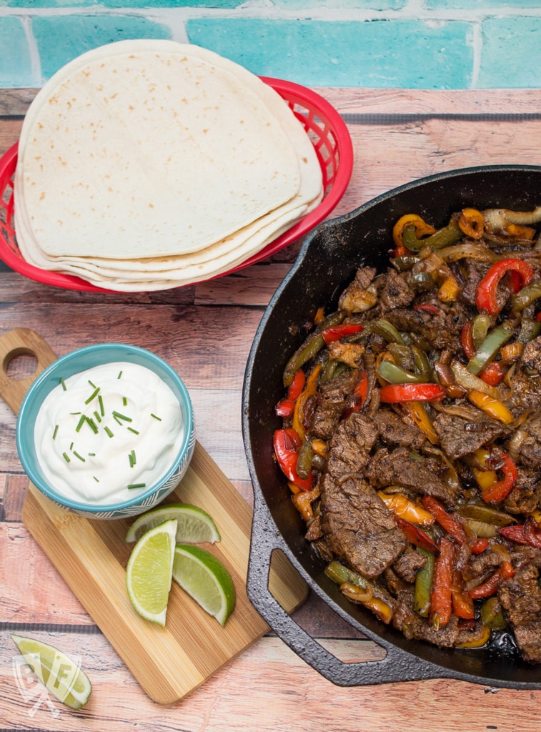 Overhead view of steak fajitas in an iron skillet with tortillas and toppings.