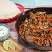 Cast iron skillet full of steak fajitas with toppings and tortillas alongside.