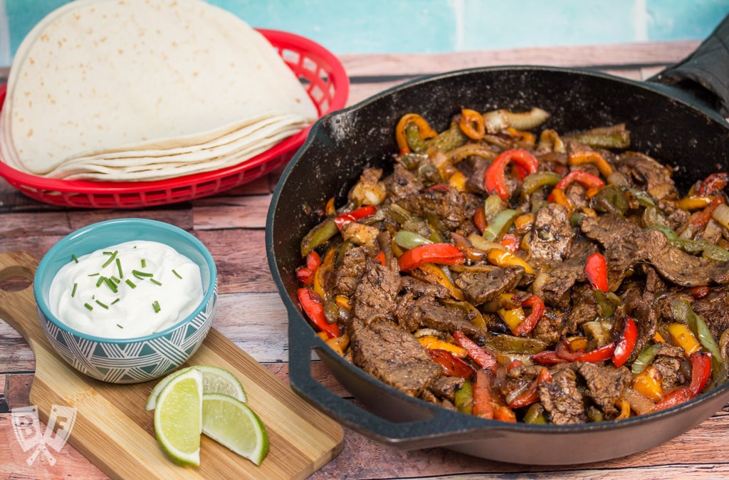 Steak fajitas in an iron skillet with toppings and tortillas alongside.
