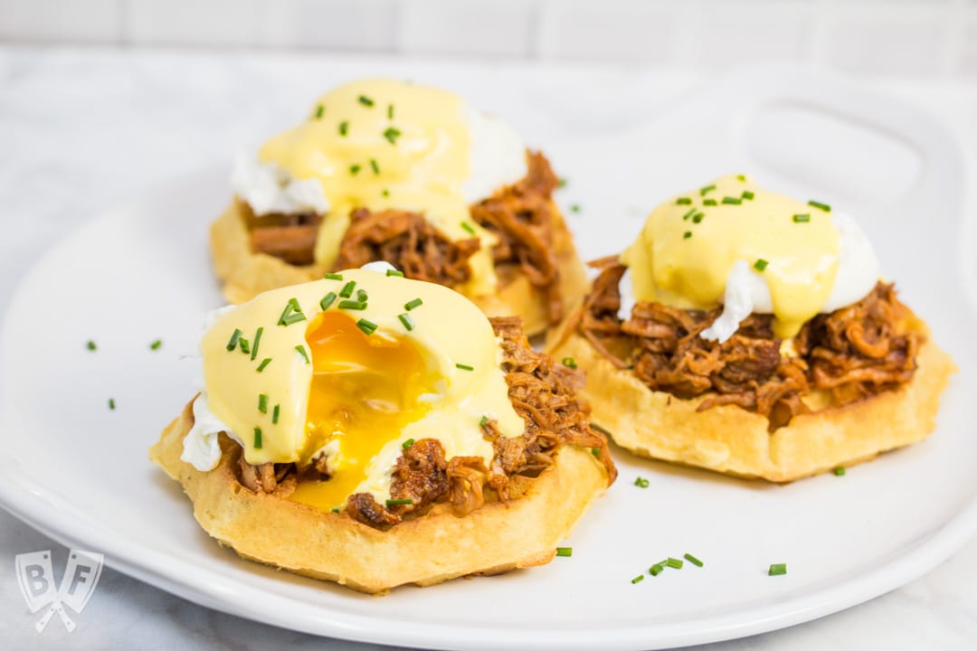A platter with 3 waffles topped with pulled pork, poached eggs, hollandaise sauce, and chives.