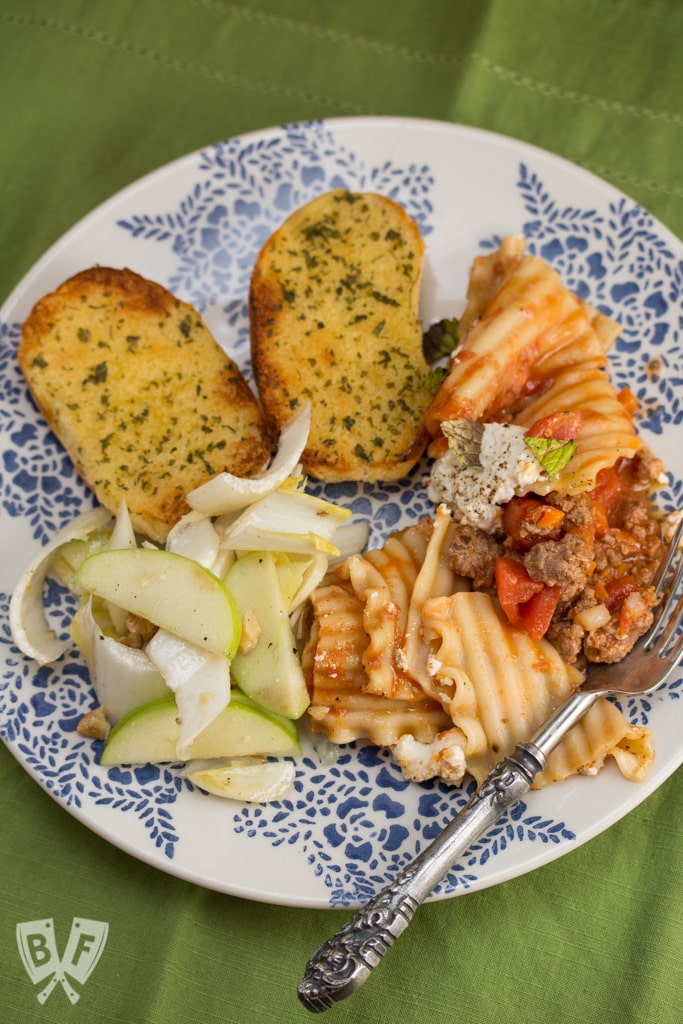 Overhead view of a plate with lamb lasagna, garlic bread, and endive salad with apples.