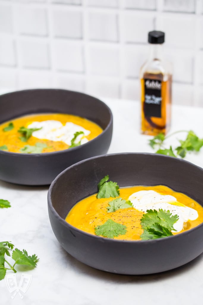 Side view of 2 bowls of curried pumpkin soup with cilantro leaves, sour cream, and chili oil garnish.