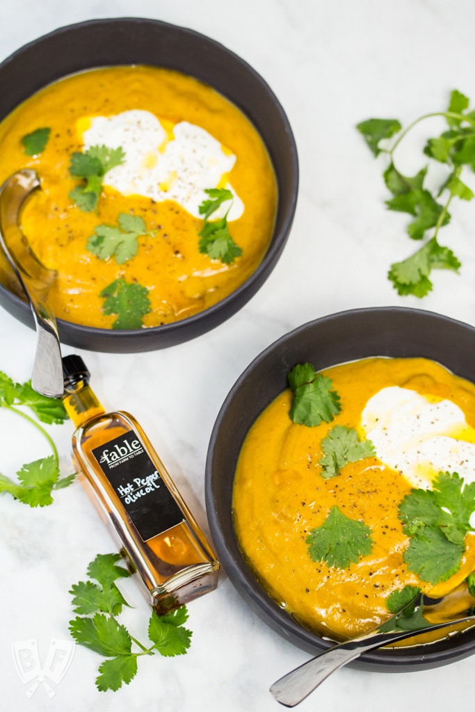 Overhead view of 2 bowls of curried pumpkin soup with cilantro leaves, sour cream, and a bottle of chili oil.