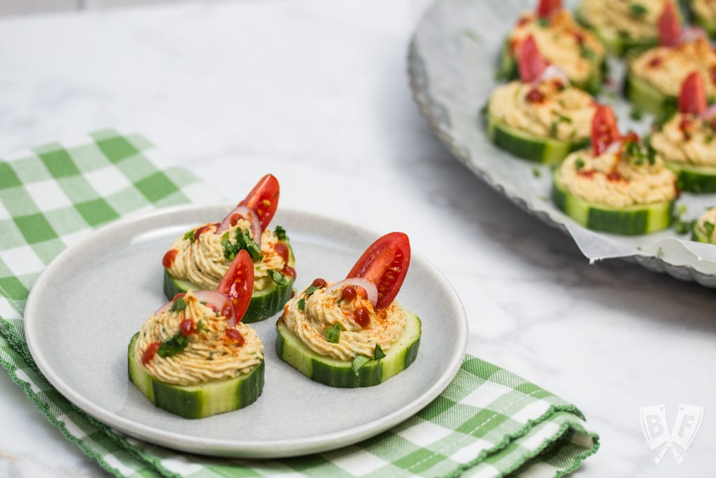 Close up of a few cucumber slices topped with a spiced chickpea purée and a platter in the background.