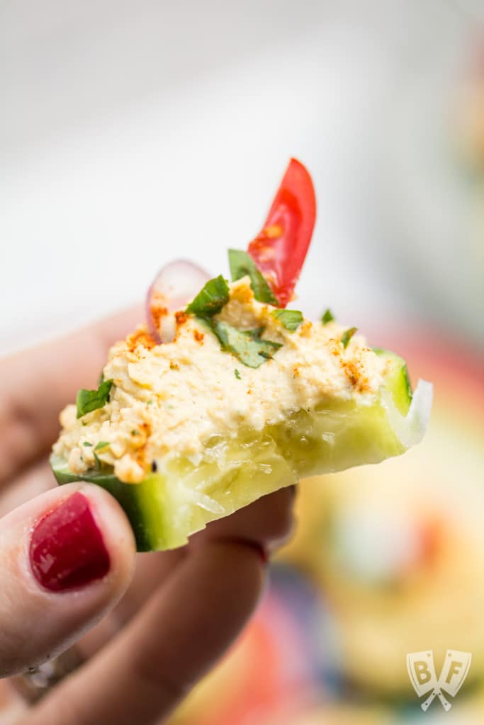 Close up of a hand holding a cucumber slice topped with a spiced chickpea purée with a bite taken out of it.