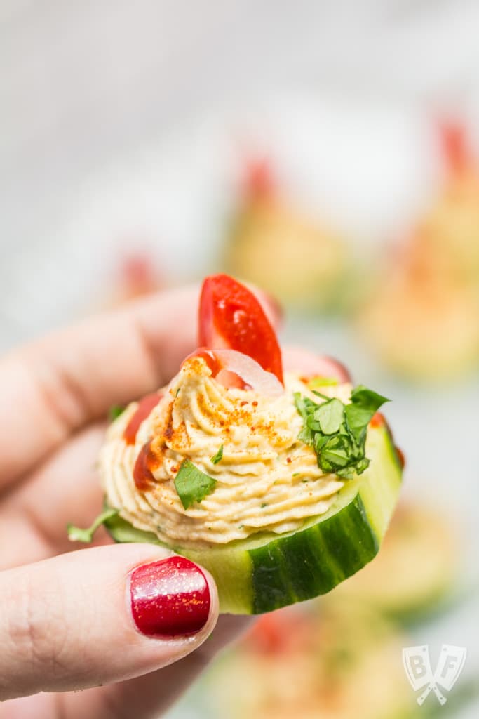 Close up of a hand holding a cucumber slice topped with a spiced chickpea purée with a platter out of focus in the background.