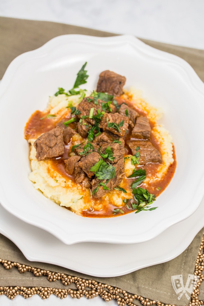 A heaping portion of mashed potatoes in a bowl with beef goulash poured over top, sprinkled with parsley.