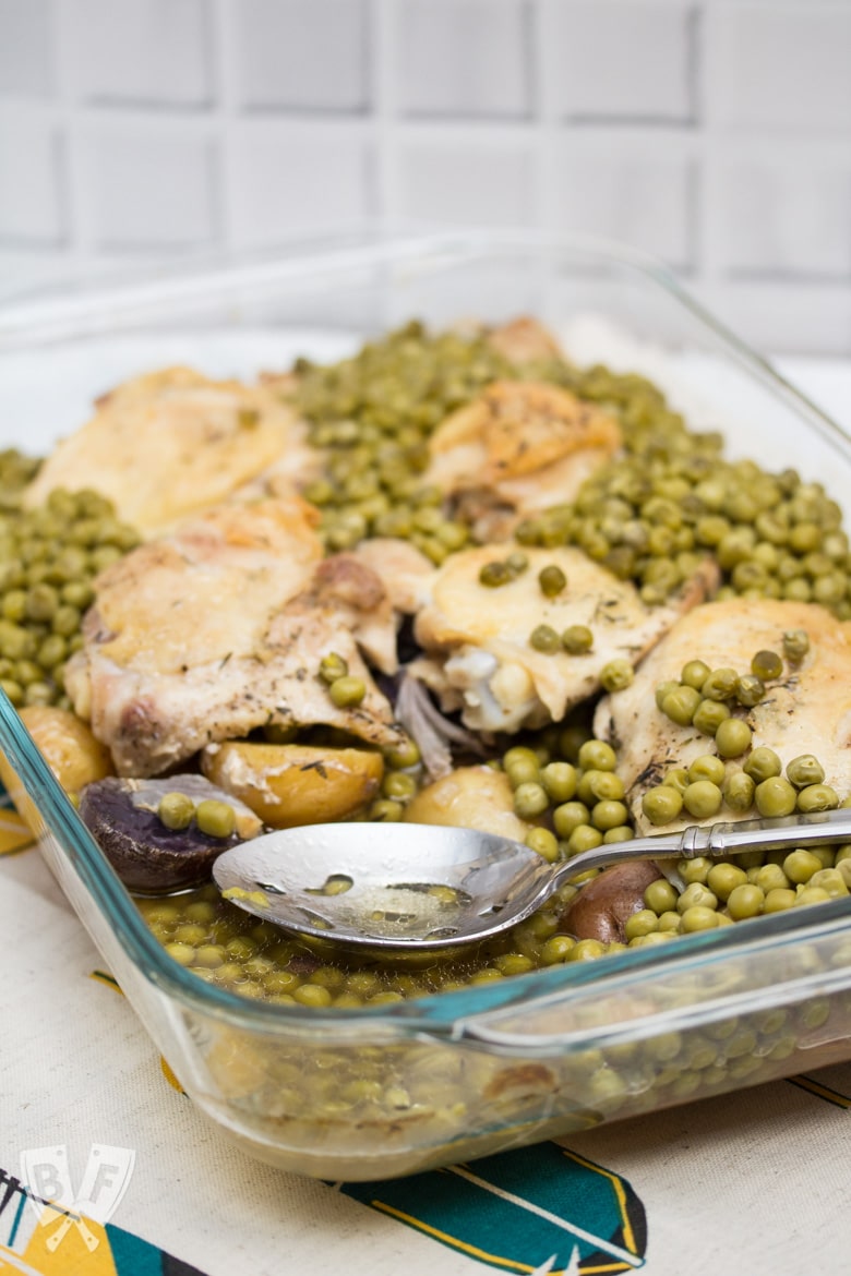 Close up of a serving spoon in a casserole dish filled with baked chicken thighs, peas, and potatoes.