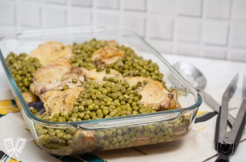 3/4 view of a casserole dish filled with baked chicken thighs, peas, and potatoes with tongs and a serving spoon.