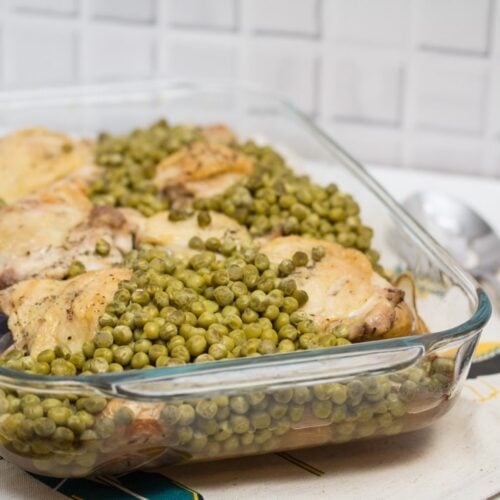 ¾ view of a casserole dish filled with baked chicken thighs, peas, and potatoes with tongs and a serving spoon.