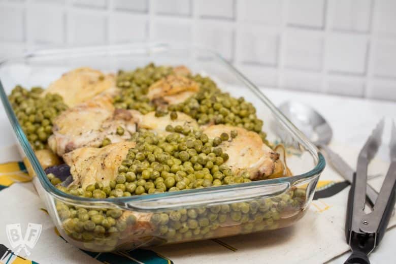 ¾ view of a casserole dish filled with baked chicken thighs, peas, and potatoes with tongs and a serving spoon.