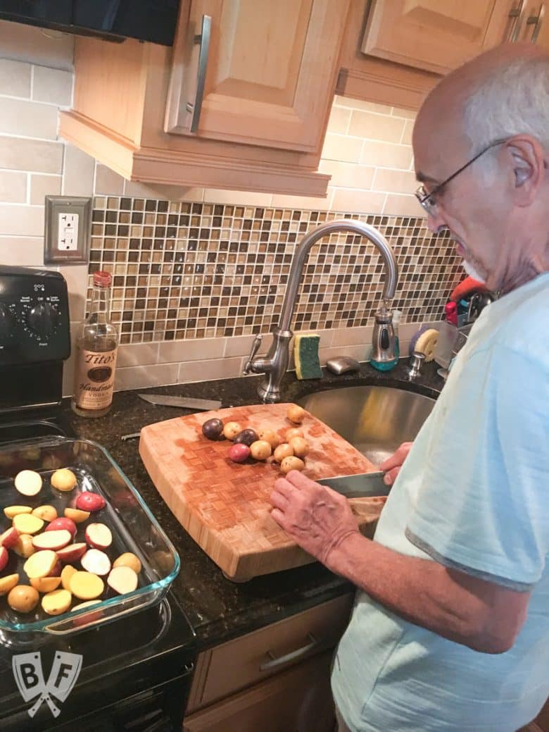 Over-the shoulder photo of a man in a kitchen cutting potatoes and placing them into a casserole dish.