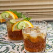 Two glasses with Pimm's cup cocktails garnished with cucumber, orange, lemon, and mint.