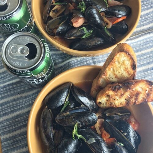 Overhead view of cooked mussels in bowls with grilled bread and cans of bear alongside.