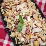 A platter of pasta salad with bits of salami, Parmesan cheese, tomatoes, and fresh basil.